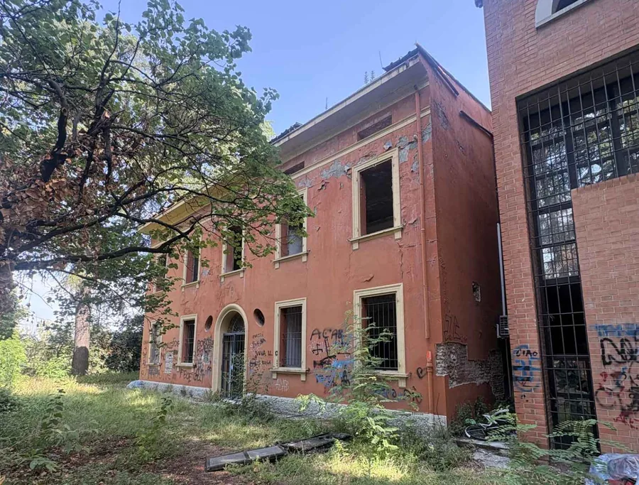 Villa A1 as found: an orange-rendered villa with barred windows, peeling render and graffiti, overgrown at its base