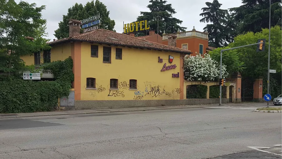 Building B as found: a yellow two-storey former hotel on Viale Lenin, its sign still up, behind a low wall and hedges